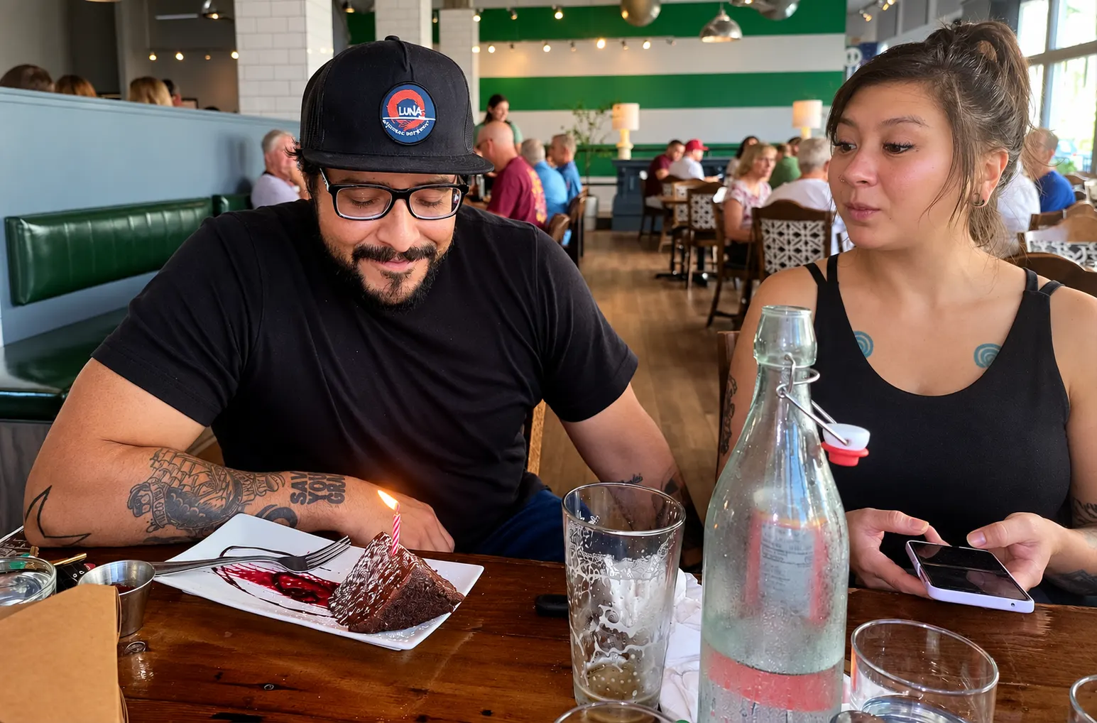 The two owners of LŪNÄ at a restaurant table, one in a LŪNÄ cap smiling at a slice of chocolate cake with a lit candle